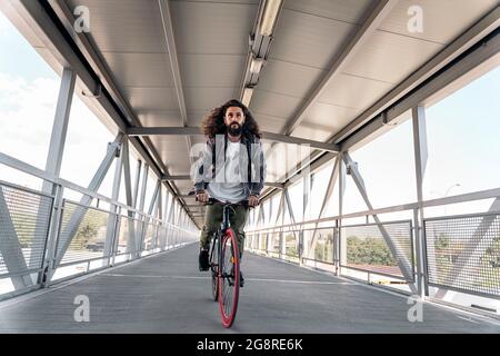 Bell'uomo hipster con capelli lunghi guardando davanti e cavalcando la sua bicicletta in città. Foto Stock