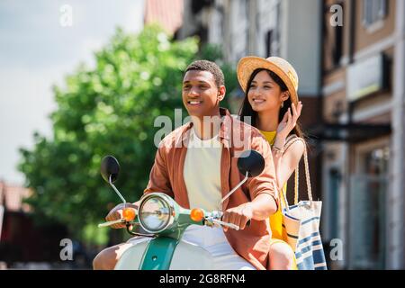 allegra donna asiatica che tocca il cappello di paglia mentre si guida scooter con il ragazzo afroamericano Foto Stock