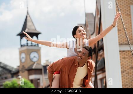 allegra donna asiatica piggybacking su felice uomo afroamericano all'aperto Foto Stock