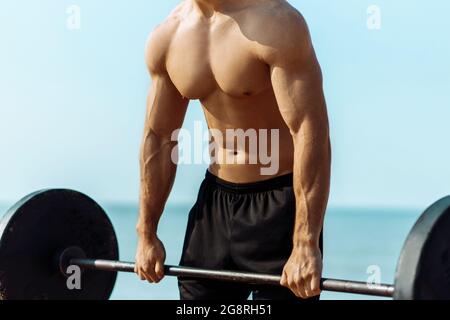 Giovane muscoloso che fa allenamento al petto con manubri di grandi pesi, sulla spiaggia all'aperto, bell'uomo con grossi muscoli, in posa Foto Stock