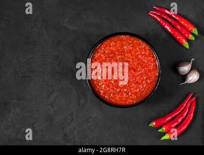 Adjika su sfondo nero. Salsa di Harissa in un recipiente con peperoncini piccanti. Spazio di copia, vista dall'alto, disposizione piatta. Foto Stock