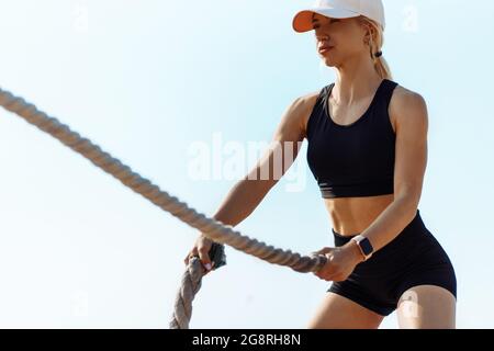 Donna fitness che usa corde di allenamento per lo sport, sulla spiaggia all'alba, atleta che si esercita con corde da combattimento, che si esercita in riva al mare, godendo il fr Foto Stock
