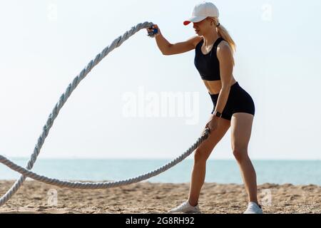 Donna fitness che usa corde di allenamento per lo sport, sulla spiaggia all'alba, atleta che si esercita con corde da combattimento, che si esercita in riva al mare, godendo il fr Foto Stock