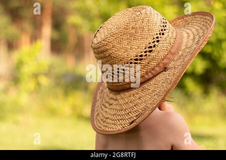 Un piccolo bambino in un cappello da fattoria di paglia guarda in lontananza con la schiena alla macchina fotografica. Meravigliosi momenti d'infanzia nella fattoria Foto Stock
