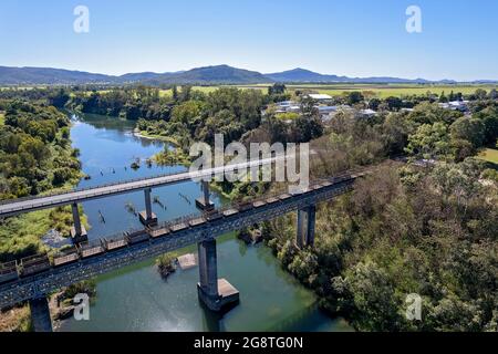 Antenna di bidoni vuoti di canna da zucchero che vengono tirati attraverso un torrente paese da un treno, parallelo ad un ponte autostradale Foto Stock