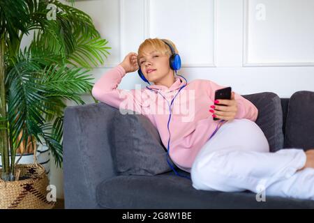 Una giovane donna si siede sul divano con un tablet e ascolta la musica attraverso le cuffie. Cultura giovanile. Foto Stock