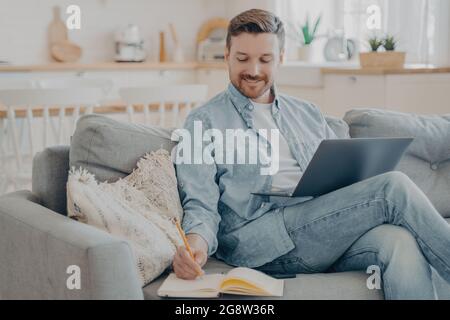 Giovane lavoratore in ufficio che lavora in remoto da casa per la ricerca utilizzando un notebook e prendendo appunti Foto Stock