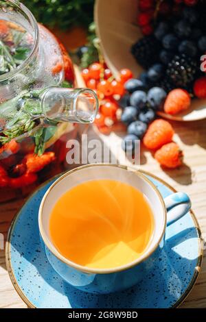 tè naturale aromatico alla frutta in una teiera trasparente e in una tazza tra frutti di bosco e fiori. tè aromatico scaldante con un profumo profondo di frutti di bosco e di bosco Foto Stock