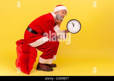 L'uomo anziano annoiato indossa il costume di babbo natale seduto sulla borsa rossa grande con i regali e tenendo l'orologio da parete nelle mani, tempo di attesa per dare il presente. Indo Foto Stock