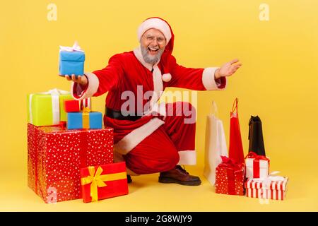 Gentile uomo anziano amichevole con la barba grigia che indossa il costume di babbo natale seduto circondato da scatole presenti e borse per la spesa, saluto con il nuovo anno. Foto Stock