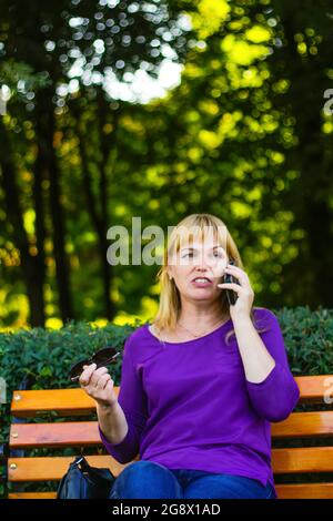 Defocus caucasica emozione bionda donna che parla, parlando al telefono esterno, all'aperto. donna di 40 anni in blusa viola nel parco. Persone adulte Foto Stock