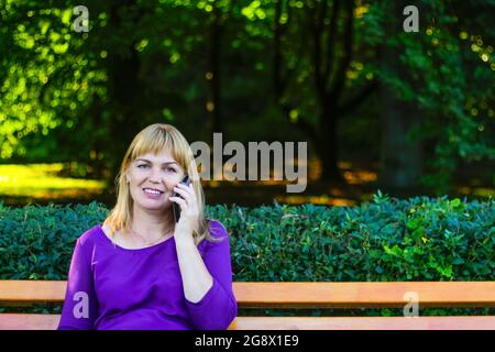 Defocus bionda caucasica sorridente donna che parla al telefono all'esterno, all'aperto. donna di 40 anni in blusa viola nel parco. Femmina adulto Foto Stock