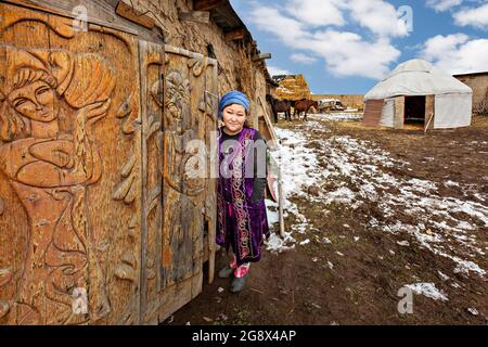 Donna kirghiza in abiti tradizionali con porta scolpita in primo piano e un yurta nomade sullo sfondo vicino Bishkek, Kirghizistan Foto Stock