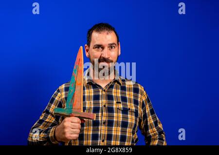 Uomo in una camicia gialla in plaid graffiando la schiena con una spada di legno isolata su sfondo blu studio. Foto Stock