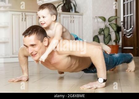 Papà e figlio fanno push-up dal pavimento nella stanza della casa. Il bambino giace sulla schiena di suo padre. Attività fisica a casa. Un buon modello di ruolo. Un uomo e un ragazzo giocano a casa. Foto Stock