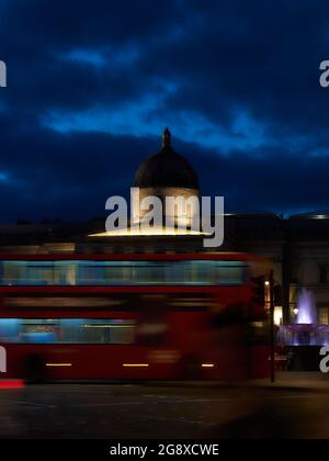 Immagine energica che mostra un autobus londinese sfocato nel movimento che passa di fronte a una National Gallery gridata di notte in Trafalgar Square. Foto Stock