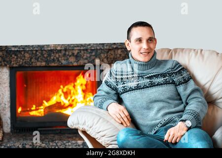 Un giovane in un maglione lavorato a maglia si siede in una sedia di pelle contro lo sfondo di un camino con fuoco e sorrisi. Comfort e calore a casa da un caminetto di lusso. Spazio copia Foto Stock