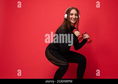 Foto a profilo laterale di bella felice sorridente giovane donna brunet che indossa abiti sportivi neri isolati sulla parete di fondo rossa squating utilizzando manubri Foto Stock