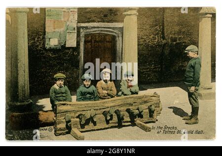 Primi anni '900 tinted saluti scheda di giovani ragazzi seduti nelle scorte, una forma antiquata di punizione attraverso l'umiliazione del criminale, anche se posato per questa foto. Totnes Old Guild Hall, Devon, Regno Unito pubblicato 1913 Foto Stock