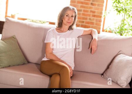 Foto di affascinante dolce vecchia donna vestita t-shirt bianca seduta divano sorridente casa casa interna piatta Foto Stock