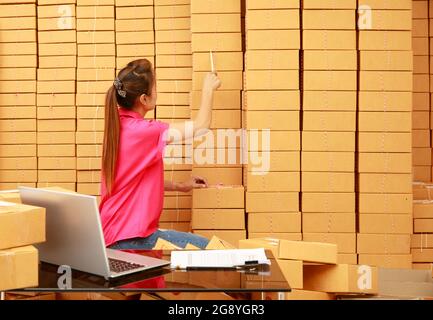 La donna asiatica usa le scatole di pacchi di conteggio della matita nel suo commercio onlind di shopping a casa con il calcolatore del laptop sulla scrivania Foto Stock