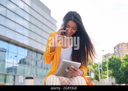 Giovane donna d'affari latina che indossa una giacca gialla che lavora con un tablet e parla sullo smartphone seduto all'aperto. Foto Stock