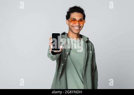 uomo afro-americano smilito mentre mostra smartphone con schermo vuoto isolato su grigio Foto Stock