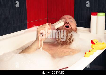 Carino uomo bearded divertente prendere bagno bolla con anatre di gomma e cantare in pennello da bagno godendosi relax a casa dopo dura giornata. Foto Stock