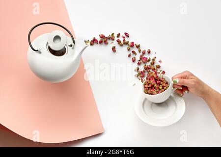 Tè secco alle erbe di fiori di rosa. Piatto surreale con fiori di rosa essiccati che versano da pentola di tè in ceramica bianca in tazza in mano femminile. Fiore di tè alle erbe Foto Stock
