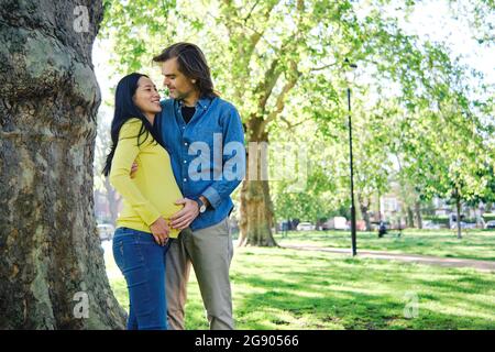 Donna incinta sorridente che guarda il marito dal tronco dell'albero Foto Stock