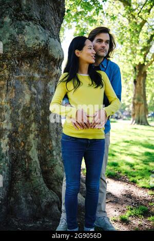 Donna incinta sorridente che guarda via mentre parla con il marito di fronte all'albero al parco Foto Stock