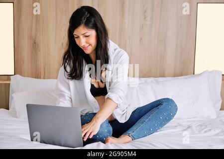 Donna incinta di adulto medio che usa un computer portatile mentre si siede sul letto in camera da letto Foto Stock