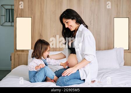 Ragazza carina che tocca il ventre delle madri incinte mentre si siede sul letto a casa Foto Stock