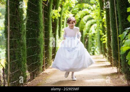 Ragazza carina in abito bianco di comunione che corre sul sentiero in giardino verde Foto Stock