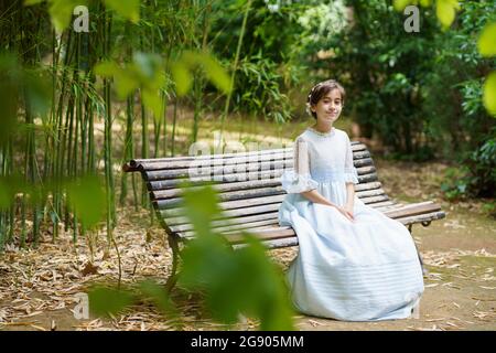 Ragazza sorridente seduta in abito bianco sulla panca del parco Foto Stock