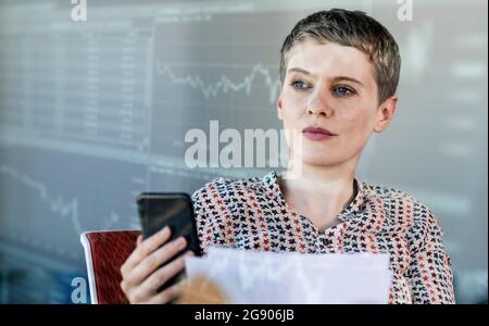 Donna professionista che tiene uno smartphone mentre guarda il grafico sulla parete digitale Foto Stock