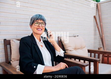 Donna d'affari matura con occhiali che parlano sullo smartphone nel cortile Foto Stock
