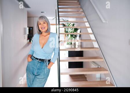 Donna matura con le mani in tasche appoggiate su ringhiera di vetro da gradini a casa Foto Stock