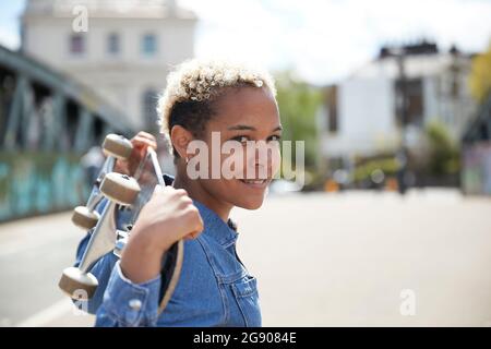 Donna sorridente che porta lo skateboard a spalla durante la giornata di sole Foto Stock