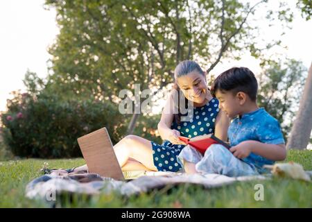 Donna sorridente che aiuta il nipote a leggere il libro di storia nel parco pubblico Foto Stock