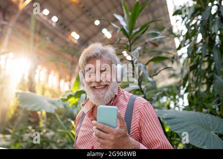 Uomo con bearded bianco felice che ascolta la musica attraverso le cuffie senza fili mentre si trova di fronte alle piante Foto Stock