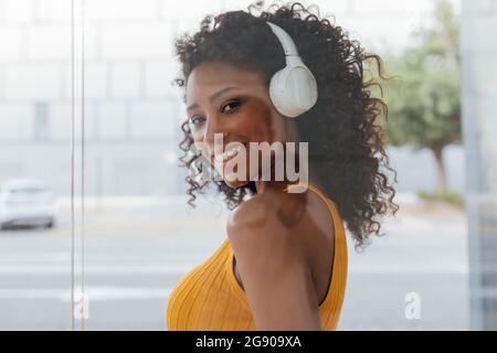 Donna sorridente con capelli ricci che ascolta la musica attraverso le cuffie wireless viste attraverso la parete di vetro Foto Stock