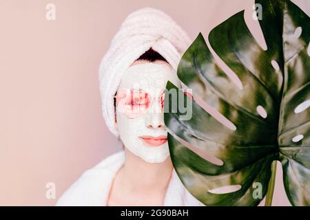 Giovane donna con maschera di bellezza che copre l'occhio con foglia monstera Foto Stock
