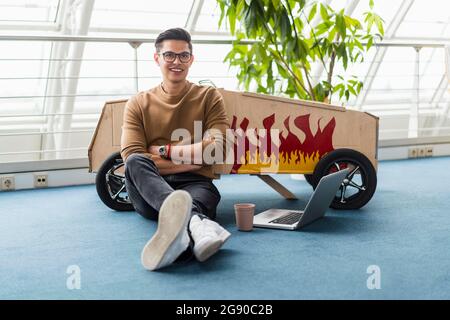 Uomo d'affari creativo sicuro con le braccia incrociate seduto da un computer portatile e un'auto giocattolo in ufficio Foto Stock