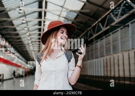 Ragazza allegra che indossa un cappello usando lo smartphone alla stazione ferroviaria Foto Stock