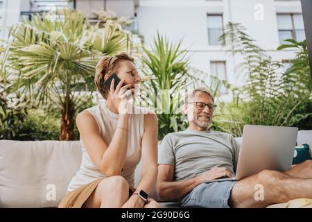 Donna che parla al telefono cellulare da uomo utilizzando un computer portatile in terrazza Foto Stock