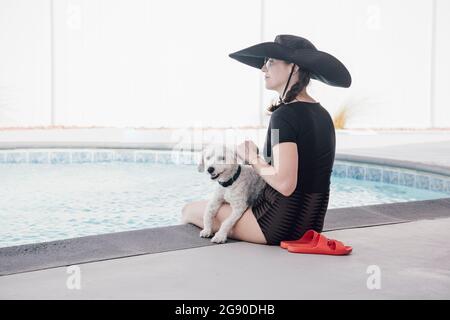 Donna in cappello che guarda via mentre si siede con il cane a bordo piscina nel patio Foto Stock