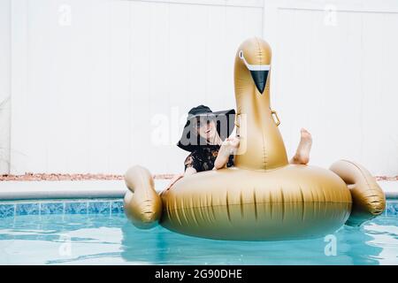 Donna felice che indossa un cappello seduto su un cigno gonfiabile in piscina Foto Stock