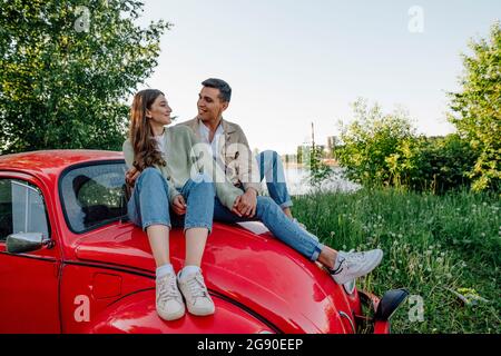 Sorridente coppia che si guarda l'un l'altro seduto su un cofano auto rosso Foto Stock