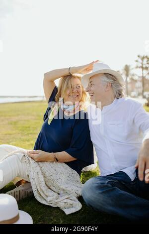 Donna matura felice guardando l'uomo che indossa il cappello mentre si siede sull'erba Foto Stock